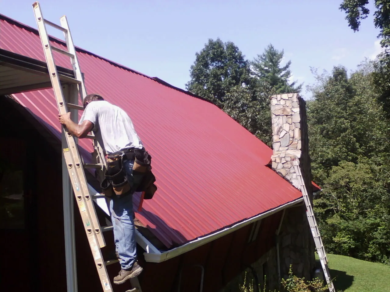Roofer on a ladder beside a red metal roof and stone chimney — Soffit & Fascia Repair by Roofing Brothers in Greenwood Springs